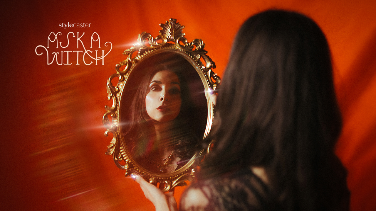 Woman holding a gold and ornate oval mirror, displaying her reflection in front of an orange backdrop.