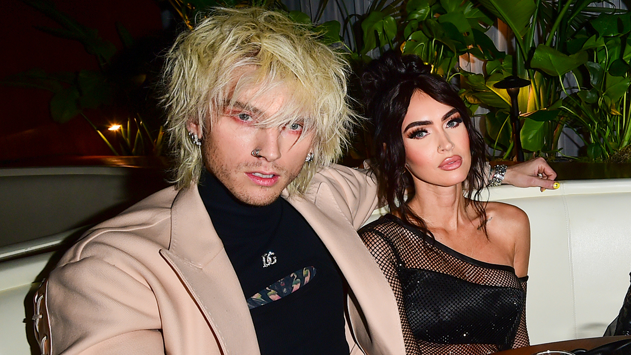 Machine Gun Kelly, a blonde white man wearing a blazer and black top posing with Megan Fox, a white woman with black hair and a black net dress.