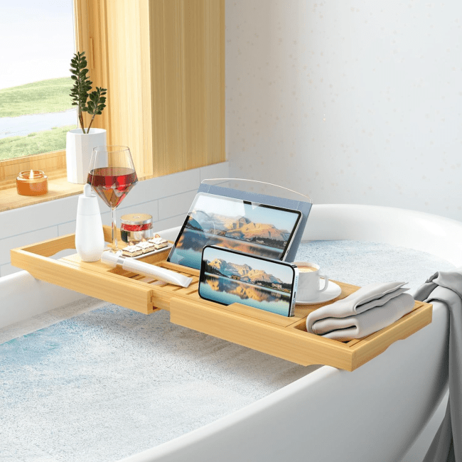 A full bathtub topped with a bath tray holding a glass of wine, a tablet, a phone, and a mug of coffee