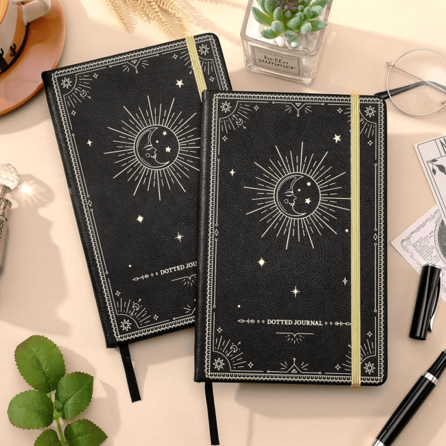 Two black leather journals decorated with gold suns, moons, and stars surrounded by plants and pens