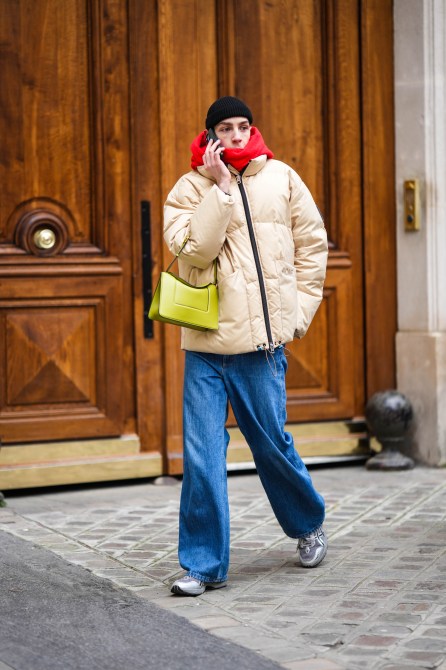 A guest wears a black wool beanie, a red hoodie sweater, a beige high neck puffer jacket, a green yellow shiny leather handbag, blue denim large pants, silver and gray leather sneakers , outside Ludovic de Saint Sernin, during Paris Fashion Week - Menswear Fall-Winter 2023-2024, on January 22, 2023 in Paris, France.