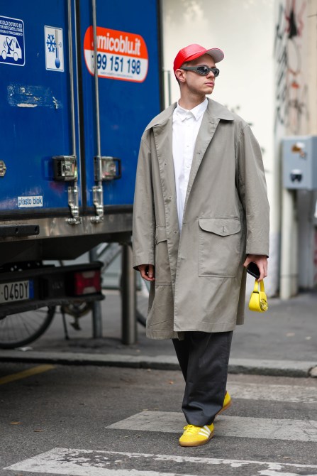 A guest wears a red vinyl cap, sunglasses, a white shirt, a beige trench coat, a tiny yellow bag, black wide-legged pants, yellow Adidas sneakers , outside Onitsuka, during the Milan Fashion Week - Womenswear Spring/Summer 2024 on September 20, 2023 in Milan, Italy.
