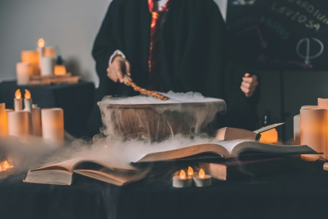 A person dressed in a Harry Potter costume holds a wand over a steaming cauldron