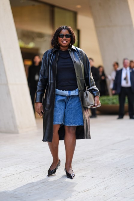 A guest wears black sunglasses, silver earrings, black t-shirt, shiny black leather trench coat, navy blue denim jean waisted rolled up shorts, dark gray leather bag, shiny black pointed toe ballerina flats, outside Toteme, during the New York Fashion week Spring/Summer 2025 on September 10, 2024 in New York, New York.