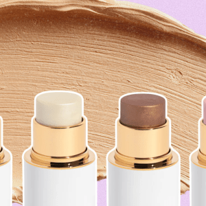 A pink background with a foundation smear and four different face sticks in blush, highlighter, and bronzer tones.